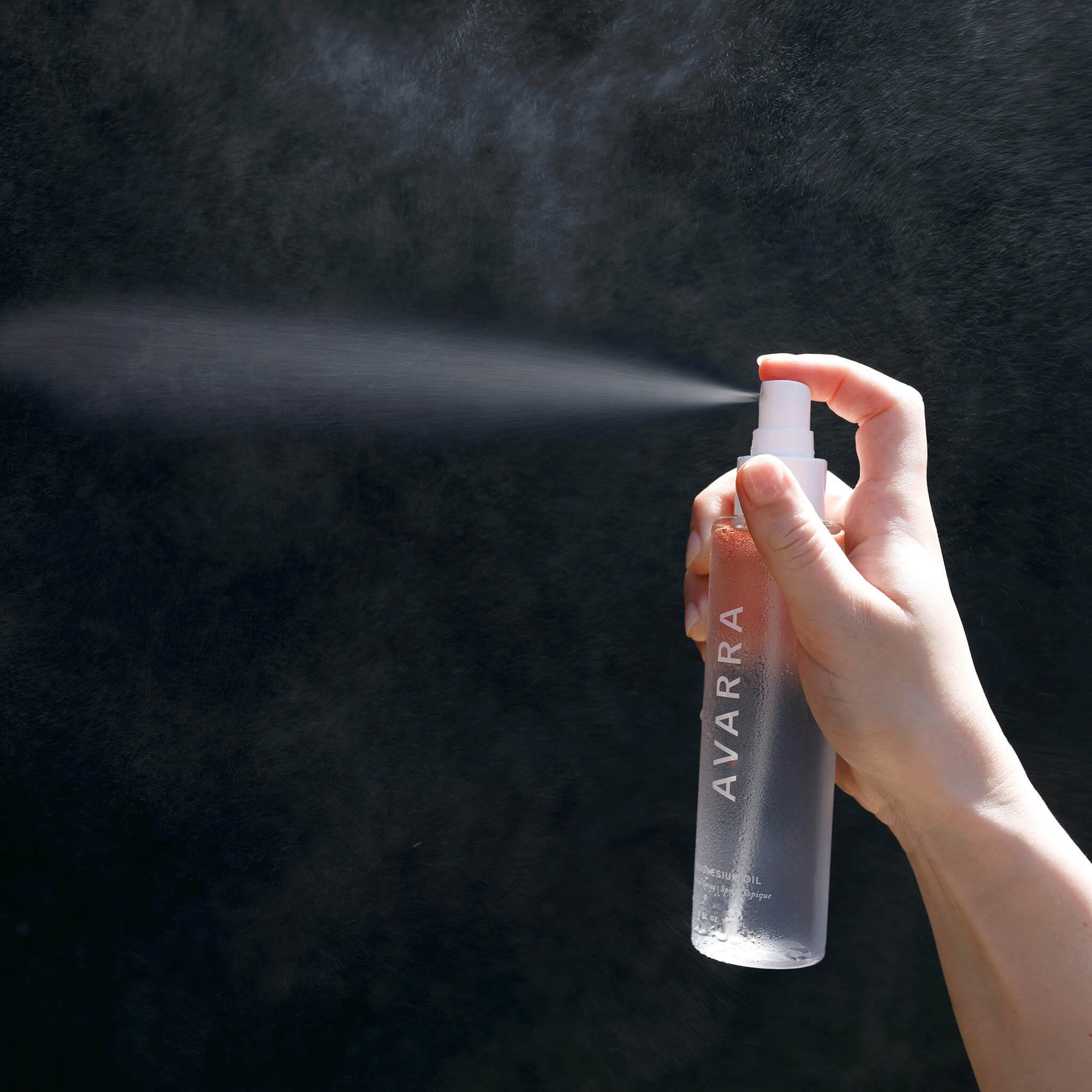 Avarra magnesium oil bottle with finger pressing the pump and a mist of spray on black background